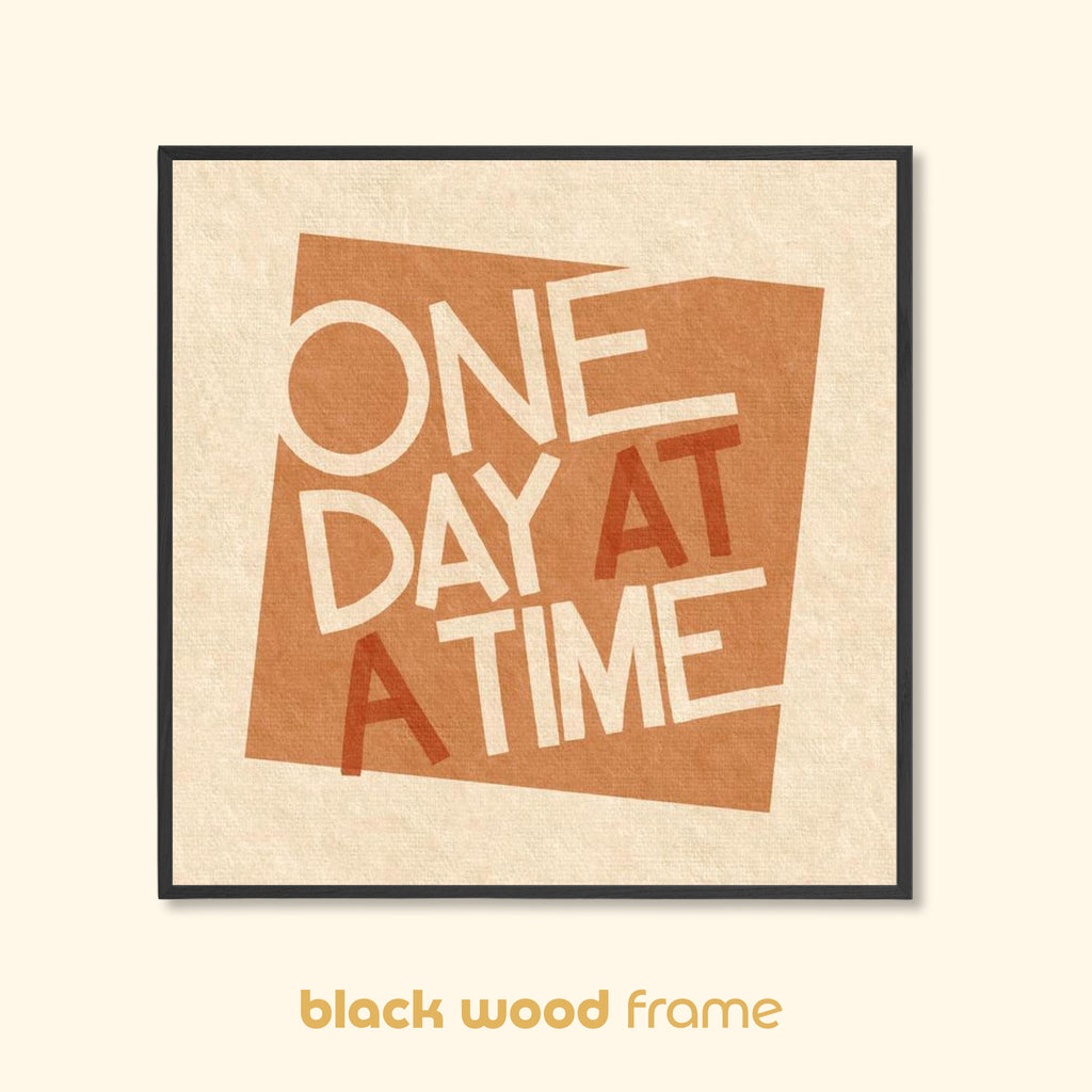 One Day at a Time typographic art print in a black wood frame, featuring warm earthy tones and mid-century inspired lettering on a textured background