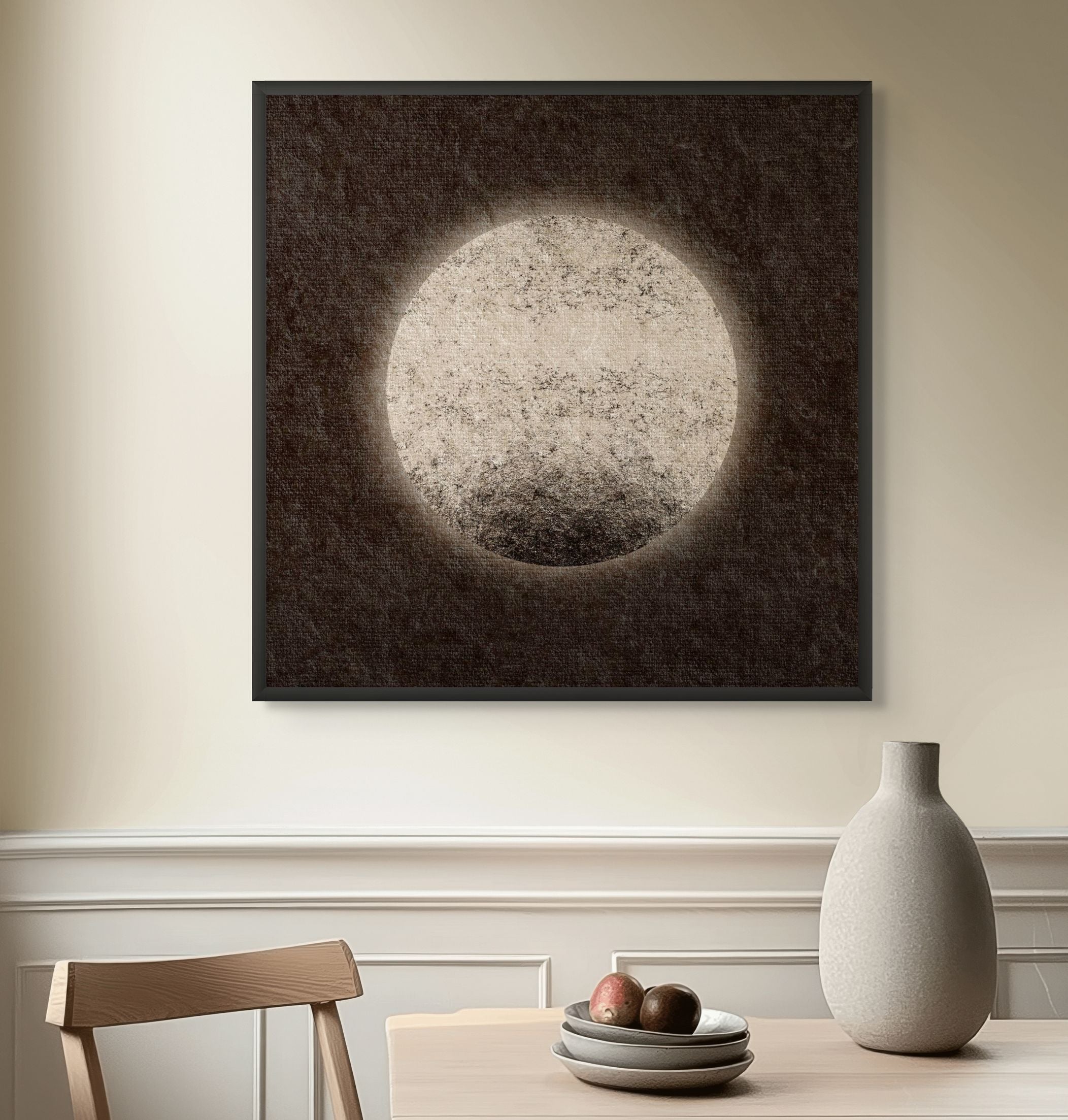 Moon-inspired abstract art print with a pale circular form and vintage textured surface, displayed in a black frame above a dining table in a calm, modern interior.